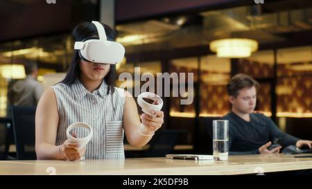 Employée de bureau asiatique utilisant un casque VR et des contrôleurs sans fil, regardant les données en réalité virtuelle. Travaillez dans un bureau moderne de la société hi-tech avec divers collègues. Technologie numérique du cyberespace Banque D'Images