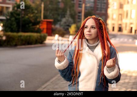 Portrait d'une jeune femme avec des dreadlocks regardant loin, touchant la coiffure colorée, debout sur la rue de la ville Banque D'Images