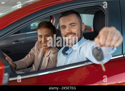 Photo d'un jeune couple assis à l'intérieur d'une nouvelle voiture. Homme lui montrant les clés. Concept de location de voiture Banque D'Images