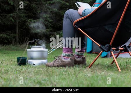 Femme lisant et faisant bouillir une bouilloire sur un poêle de camping. Banque D'Images
