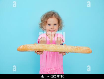 Belle petite fille montre baguette française sur fond bleu vide, espace libre de copie. Portrait d'un enfant souriant de l'âge de la maternelle tenant dans tendu Banque D'Images