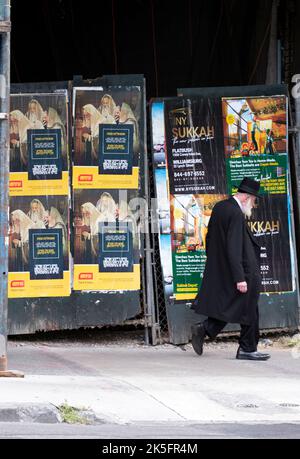 Un juif hassidique plus âgé passe devant un mur avec des affiches en yiddish et en anglais provenant de sources publicitaires pour des sucos et défendant la religion. Banque D'Images