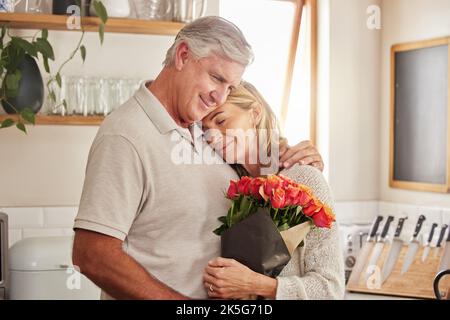 Bouquet de fleurs, câlin et couple senior pour célébrer l'amour, le mariage et l'anniversaire dans leur maison. Joyeux homme âgé et femme embrassant avec la rose Banque D'Images