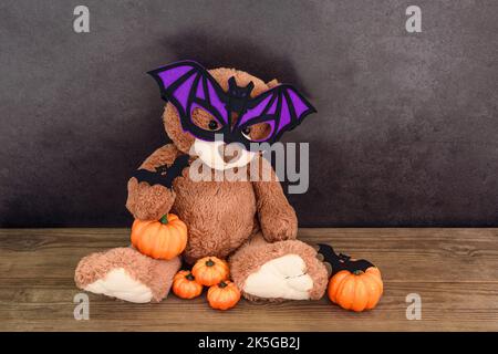 Ours en peluche brun drôle avec masque de chauve-souris et citrouilles sur une vieille table en bois au mur foncé Banque D'Images