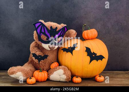 Ours en peluche brun drôle avec masque en forme de chauve-souris et citrouilles sur une vieille table en bois au mur noir Banque D'Images