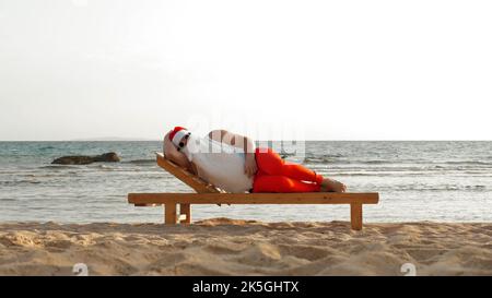 le père noël, en lunettes de soleil, se trouve sur une chaise longue en bois sur la plage, au bord de la mer. Il dort et se détend. le père noël est en vacances d'été, au bord de la mer. Photo de haute qualité Banque D'Images