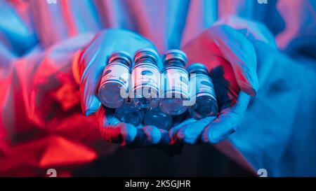 Vaccin Covid-19. Inoculation du coronavirus. Recherche en laboratoire. Soins médicaux. Flacon flacons de dose en femme médecin mains en gants ppe en rouge bleu néon lumière o Banque D'Images