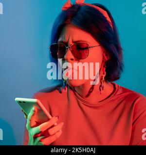 Harcèlement des noteurs. Spam en ligne. Internet faible. Une femme agacée qui colle la langue au téléphone dans un néon rouge isolé sur fond bleu. Banque D'Images