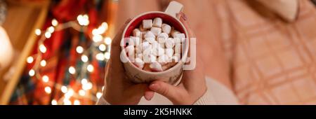 tasse de chocolat chaud ou de boisson au café avec guimauves dans la femme tenant les mains sur les jambes de la femme en pull relaxant et plaisir de l'holida Banque D'Images