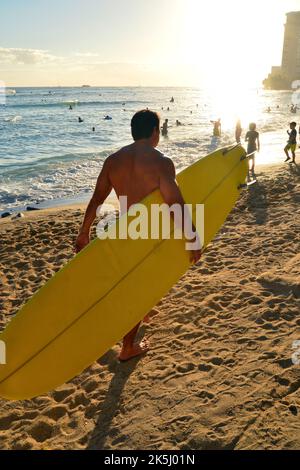 Un homme d'âge moyen transporte sa planche de surf jusqu'au rivage et les vagues lors d'une journée de vacances d'été ensoleillée à Waikiki Beach à Hawaï Banque D'Images