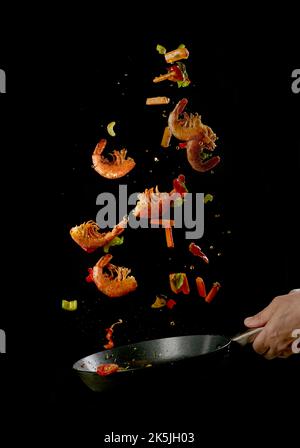 Croiser un chef anonyme qui lance des aliments dans une poêle tout en préparant de délicieuses crevettes et légumes sur fond noir Banque D'Images