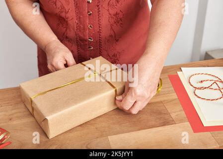 Femme adulte non reconnaissable qui enveloppe un cadeau de Noël avec du papier neutre et un ruban décoratif doré. Banque D'Images