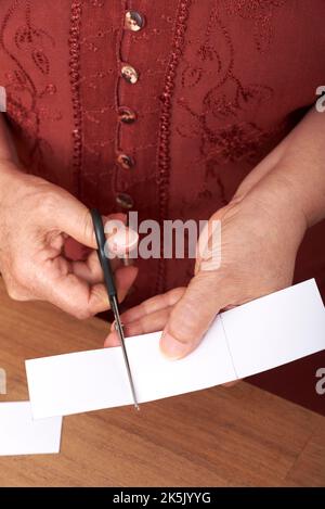 Femme mûre méconnaissable coupant avec des ciseaux de carton blanc, faisant des cartes faites à la main pour mettre ses cadeaux de Noël. Conception verticale en gros plan. Banque D'Images