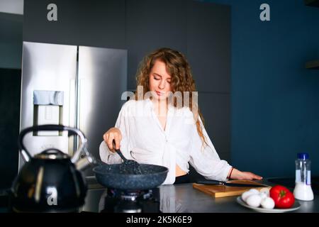 Jeune femme caucasienne souriante cuisinait le petit-déjeuner sur une poêle à frire. Jeune femme adulte faisant une omelette sur un poêle à gaz. Fille préparant son repas sur un Banque D'Images