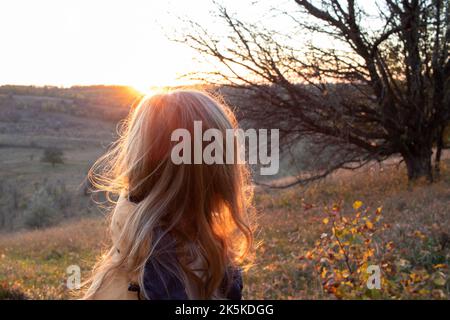 Une jeune femme aux longs cheveux blonds profite du coucher du soleil et du paysage d'automne. Vue arrière Banque D'Images