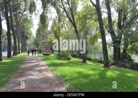 Lecco, Italie - 1 octobre 2022: Paysage avec un large trottoir de sable dans un parc vert de la ville avec des cyclistes pédalant au loin. Piste cyclable Lecco-Milan. Banque D'Images