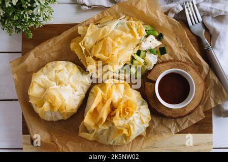 Porte-monnaie phyllo. Pâte filo remplie de poulet et de légumes sur papier parchemin et sauce barbecue. Trois sachets de pâtisseries veggie fourrées au four. Vege mixte Banque D'Images