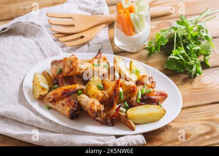 Les ailes de poulet frites dans la sauce se trouvent sur une assiette avec de grandes tranches de pommes de terre, saupoudrées d'oignons verts frais hachés. Banque D'Images
