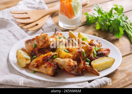 Les ailes de poulet frites dans la sauce se trouvent sur une assiette avec de grandes tranches de pommes de terre, saupoudrées d'oignons verts frais hachés. Banque D'Images