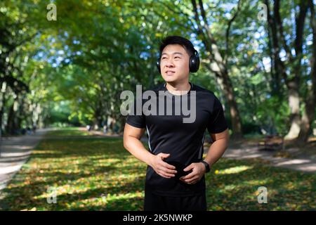 un sportif asiatique se présente le matin, un homme équipé d'un casque pour écouter de la musique et des livres audio avec des podcasts, un sportif dans un parc d'automne qui fonctionne sous le soleil. Banque D'Images