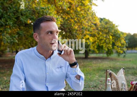 Un jeune homme souriant dans la trentaine pendant qu'il pique-niquer dans le parc passe un appel téléphonique. Banque D'Images
