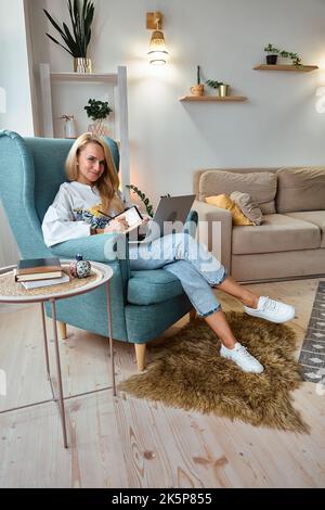 Jeune femme utilisant un ordinateur portable et écrit dans un ordinateur portable assis dans un fauteuil, travail à distance à la maison Banque D'Images