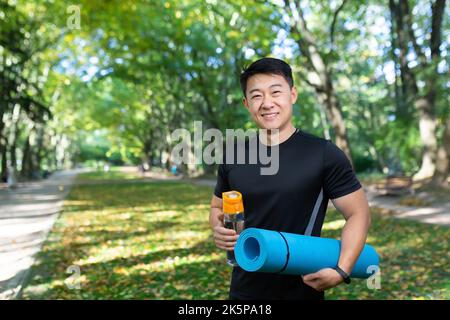 Entraîneur de fitness regardant l'appareil photo et souriant, instructeur de sport asiatique avec tapis de sport et bouteille d'eau, dans le parc d'automne le jour ensoleillé près des arbres. Banque D'Images