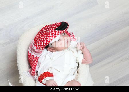 Saoudien musulman 1 mois nouveau-né garçon dans une vinaigrette arabe traditionnelle blanc thobe rouge shemagh noir agal assis sur une petite chaise douce culture de petites chaussures Banque D'Images