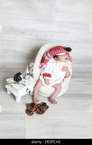 Saoudien musulman 1 mois nouveau-né garçon dans une vinaigrette arabe traditionnelle blanc thobe rouge shemagh noir agal assis sur une petite chaise douce culture de petites chaussures Banque D'Images