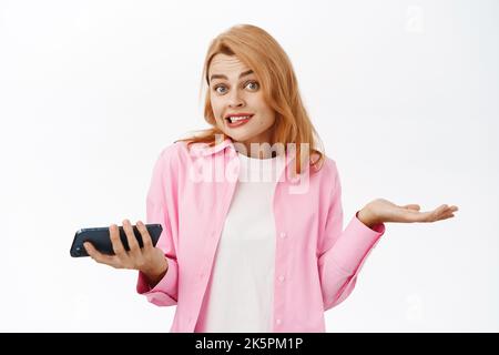 Oups, je ne sais pas. Une femme sans indice haussant les épaules avec un téléphone portable, en regardant désolé, debout sur fond blanc Banque D'Images