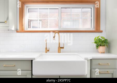 Un évier de cuisine avec un robinet en or, un évier de ferme, un fond de carreaux de métro et un meuble vert clair. Banque D'Images