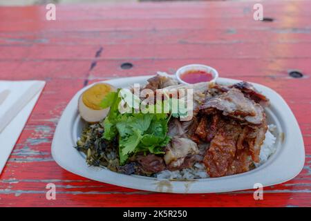 Cuisse de porc et œuf compotés avec divers légumes et riz cuit à la vapeur, sur un plat en papier et une table en bois rugueux. Banque D'Images