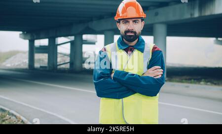 Portrait d'un jeune contremaître confiant debout à l'extérieur, portant un gilet et un casque uniformes avec un chantier de construction en arrière-plan. Concept d'entreprise de construction et de personnes. Banque D'Images