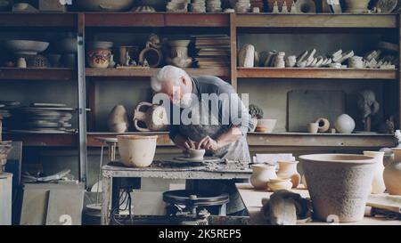 Ceramist masculin expérimenté crée des ustensiles à partir de l'argile sur la roue de potter dans l'atelier. Production de faïence, d'ustensiles faits à la main et de poterie professionnelle. Banque D'Images