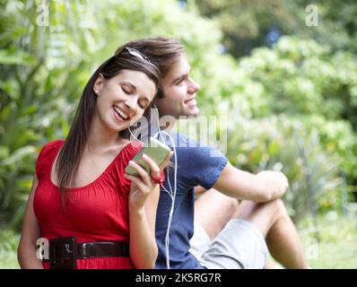 Partager un moment musical. Un jeune couple partage une paire d'écouteurs dans le parc. Banque D'Images