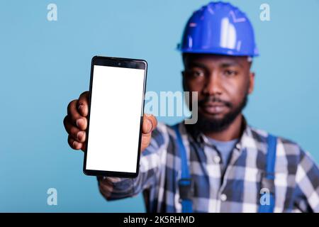 Travailleur de la construction afro-américain tenant le smartphone avec la main sur fond bleu isolé. Appareil mobile en premier plan avec écran vierge pour la publicité, prise de vue en studio. Banque D'Images