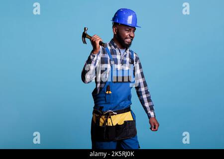 Un carrossier agressif tenant un marteau ferme un casque de protection bleu et uniforme. Afro-américain homme au travail à l'air furieux a souligné après de nombreuses heures de travail lourd sans repos. Banque D'Images