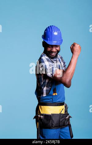 Homme fort fléchissant les muscles du bras, se préparant à travailler sur la réforme de la construction avec des outils. Un constructeur afro-américain plein d'énergie et plein d'énergie, qui pointe des biceps et des triceps, filmée en studio sur fond bleu. Banque D'Images