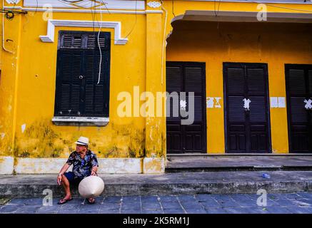 Un vieil homme vietnamien se trouve en face d'un vieux bâtiment de couleur jaune dans l'ancienne ville de Hoi an, au Vietnam. Banque D'Images