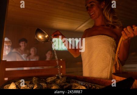 Bonne femme tenant le seau et la cuillère versant de l'eau sur pierre chaude dans la salle de sauna avec un groupe de personnes. Faites de la vapeur sur les pierres, le spa et le bien-être Banque D'Images