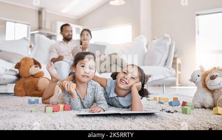 Deux petites filles dessin avec des crayons de couleur couché sur le sol de la salle de séjour avec leurs parents se détendant sur le canapé. Petits enfants sœurs frères et sœurs Banque D'Images