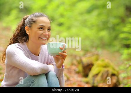 Une femme heureuse assise dans une forêt buvant un café en regardant loin Banque D'Images