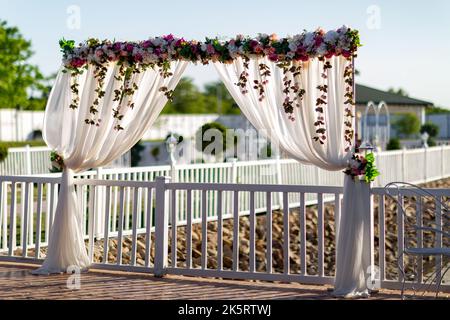 Une belle arche de mariage décorée de tissu blanc et de fleurs colorées sur la clôture en bois du parc Banque D'Images