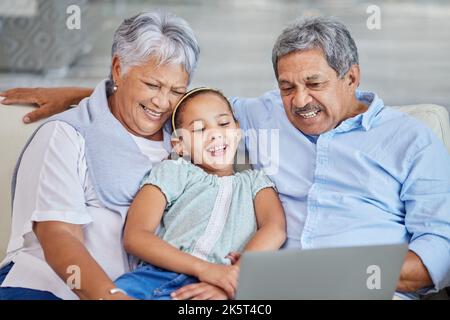 Gros plan d'un couple senior de race mixte affectueux se détendant dans leur salon avec leur petite-fille à l'aide d'un ordinateur portable. Homme et femme hispaniques Banque D'Images