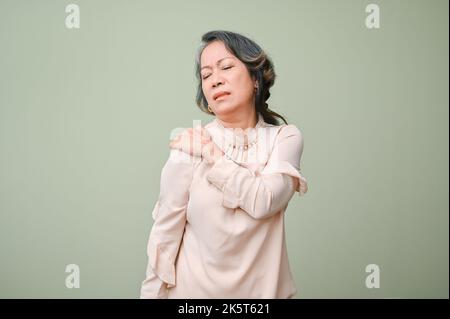 60s femme asiatique âgée souffrant de ses douleurs à l'épaule, massant son épaule, debout sur un arrière-plan isolé de studio vert. Banque D'Images