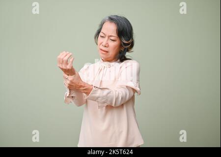 Une femme asiatique âgée, malheureuse et affligée, qui tient son poignet, souffre de douleurs sévères dans la main, massante son poignet douloureux, debout contre un g Banque D'Images