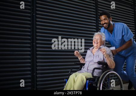 Homme soignant poussant une femme âgée excitée en fauteuil roulant dans la rue, en appréciant le temps ensemble. Banque D'Images