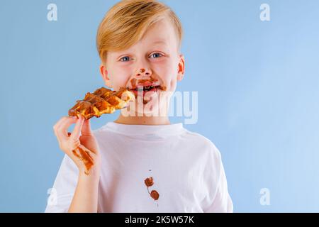 L'enfant mange de la gaufre belge avec de la sauce au chocolat sur fond bleu. Taches de chocolat sales sur les vêtements et les mains Banque D'Images