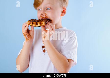 L'enfant mange de la gaufre belge avec de la sauce au chocolat sur fond bleu. Taches de chocolat sales sur les vêtements et les mains Banque D'Images