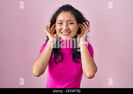 Jeune femme asiatique debout sur fond rose souriant tirant les oreilles avec les doigts, geste drôle. Problème d'audition Banque D'Images